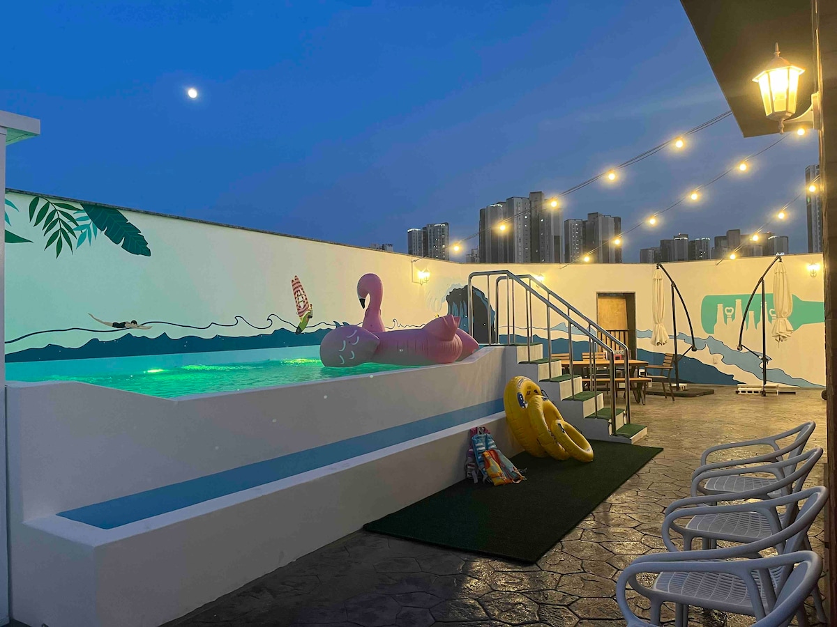 A rooftop area features a swimming pool illuminated under soft lighting. Colorful pool floats rest nearby, and an outdoor seating area is arranged with tables and chairs. A mural with tropical imagery decorates the wall, adding a vibrant touch to the space.