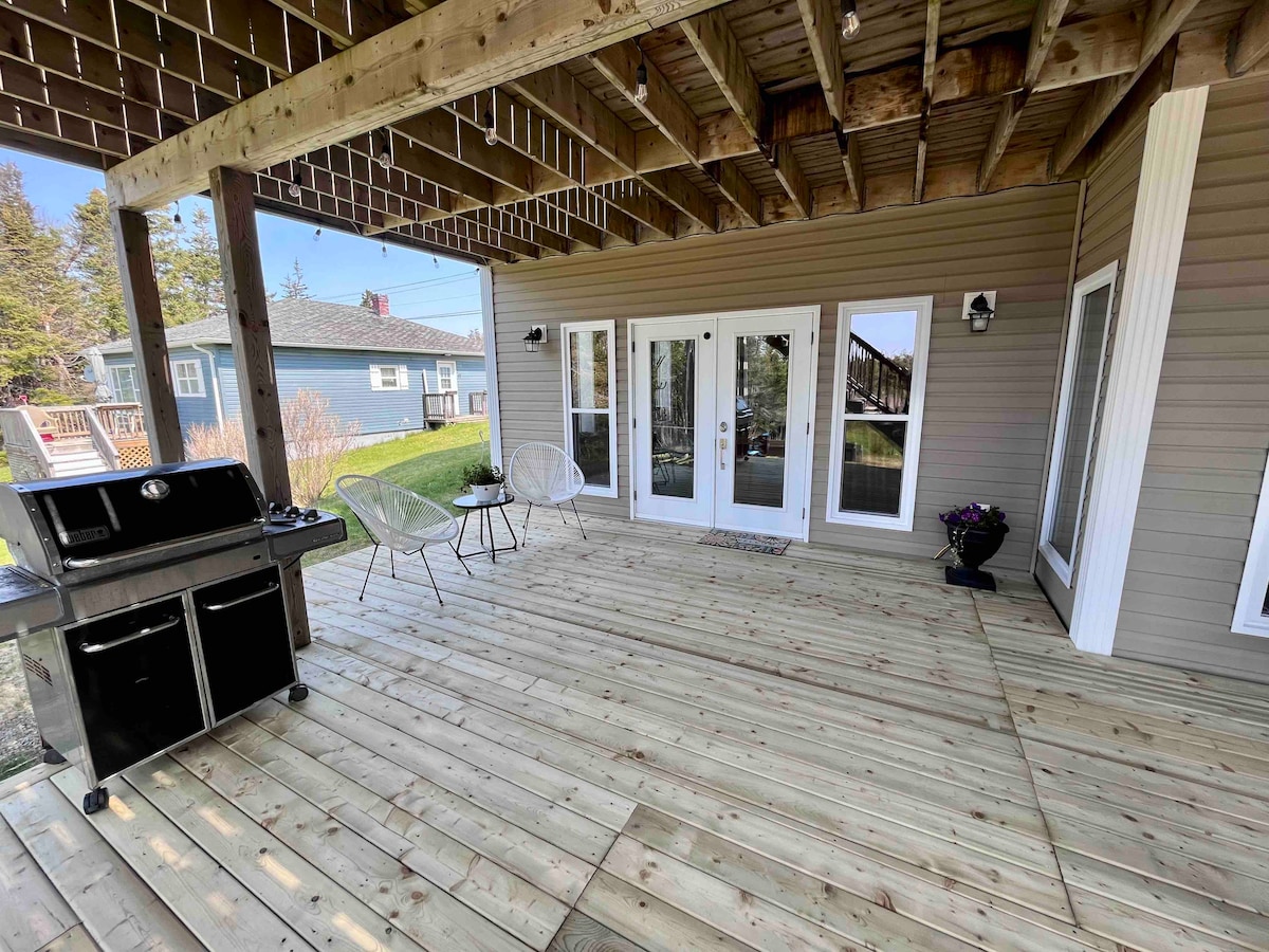 A spacious outdoor patio features wooden flooring and is partially covered, providing shade. A modern grill is situated to the left, accompanied by two stylish chairs. Glass doors lead to the interior space, and a lawn is visible in the background.