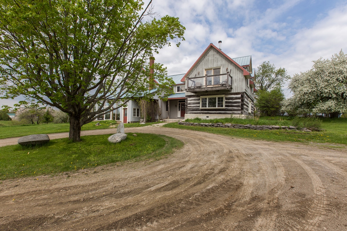 A rural driveway leads to a two-story house constructed from wood with a metal roof. A spacious front porch is visible above the entrance, surrounded by green grass and trees. The sky features clouds, hinting at a sunny day.
