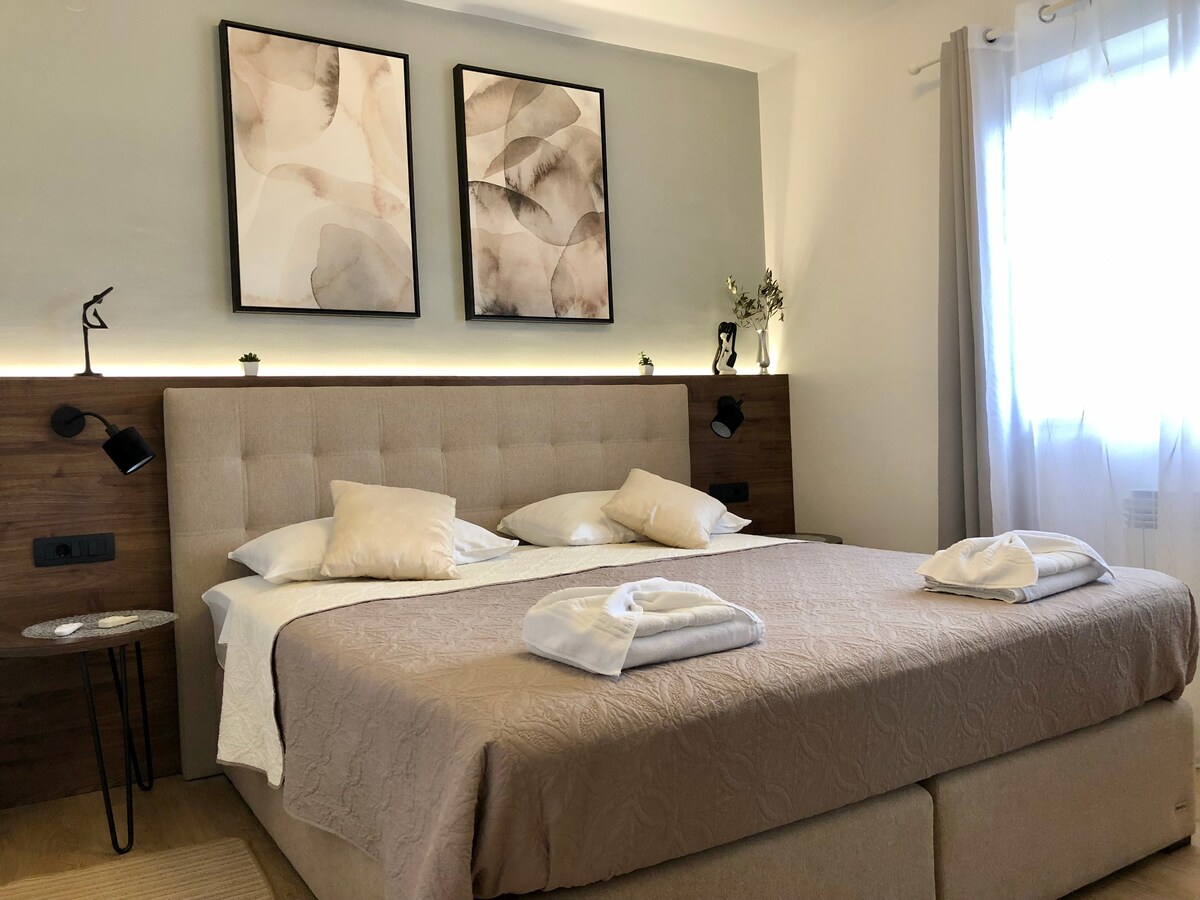 A spacious sleeping area features a large bed with multiple pillows. Two abstract artworks adorn the walls above the bed. Soft lighting is provided by wall-mounted fixtures, and a small table is positioned beside the bed. Natural light enters through a window covered by sheer curtains.