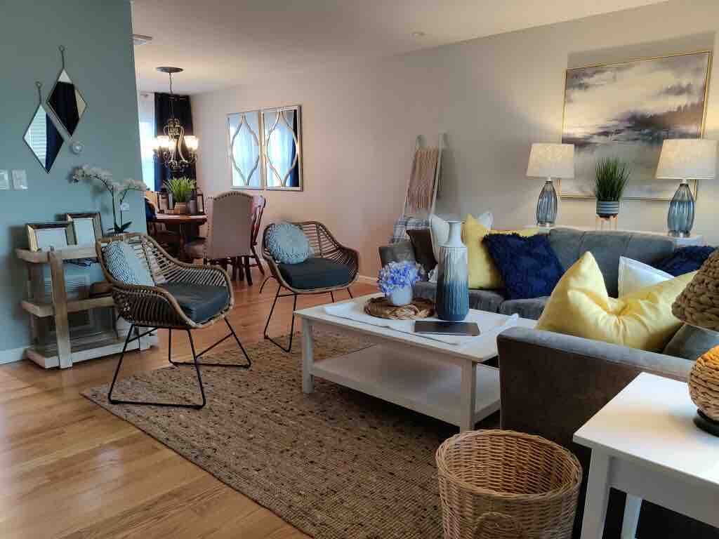A welcoming living area is featured with comfortable seating arrangements, including two wicker chairs and a spacious gray sofa adorned with decorative pillows. A coffee table sits at the center, complemented by a textured area rug. Light filters in through large windows, enhancing the overall brightness.