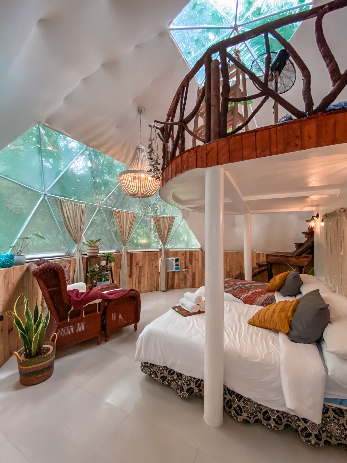 A bohemian-themed dome interior features a cozy sleeping area with a spacious bed adorned with colorful cushions. Natural light filters in through large glass panels, illuminating the seating area beside the bed. A wooden staircase leads to a lofted space, enhancing the open and airy feel.