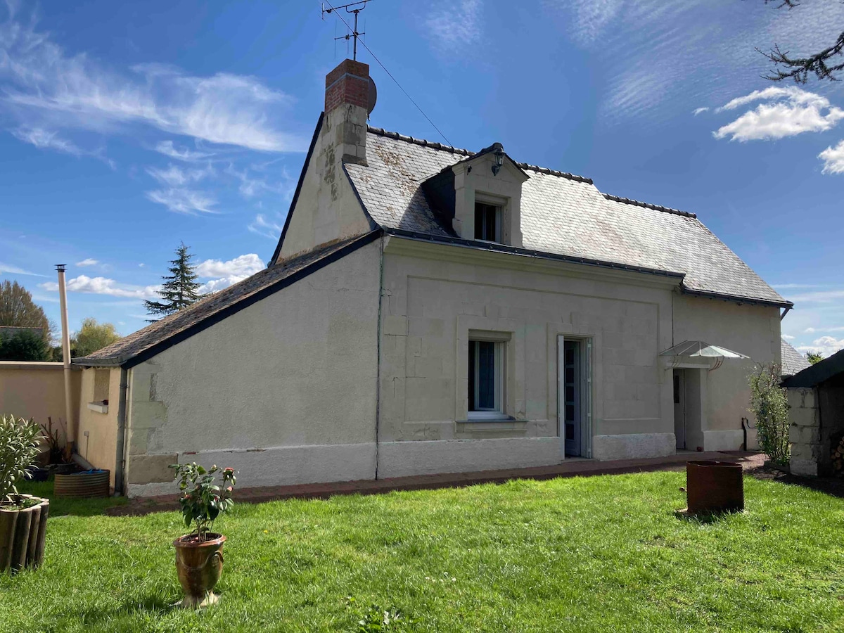 The exterior of a charming cottage is visible, featuring a sloped roof and a chimney. A spacious lawn surrounds the cottage, with a small patio area at the entrance. The walls are painted in muted tones, and windows allow natural light to enter the interior.