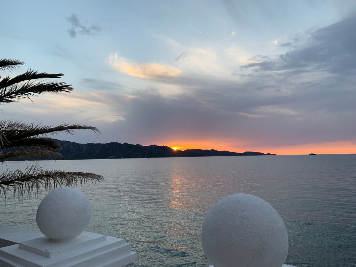 The image captures a serene sunset over calm waters, with hues of orange and purple blending in the sky. Silhouetted mountains provide a backdrop, while two white spherical structures frame the foreground, complemented by the branches of a nearby palm tree.