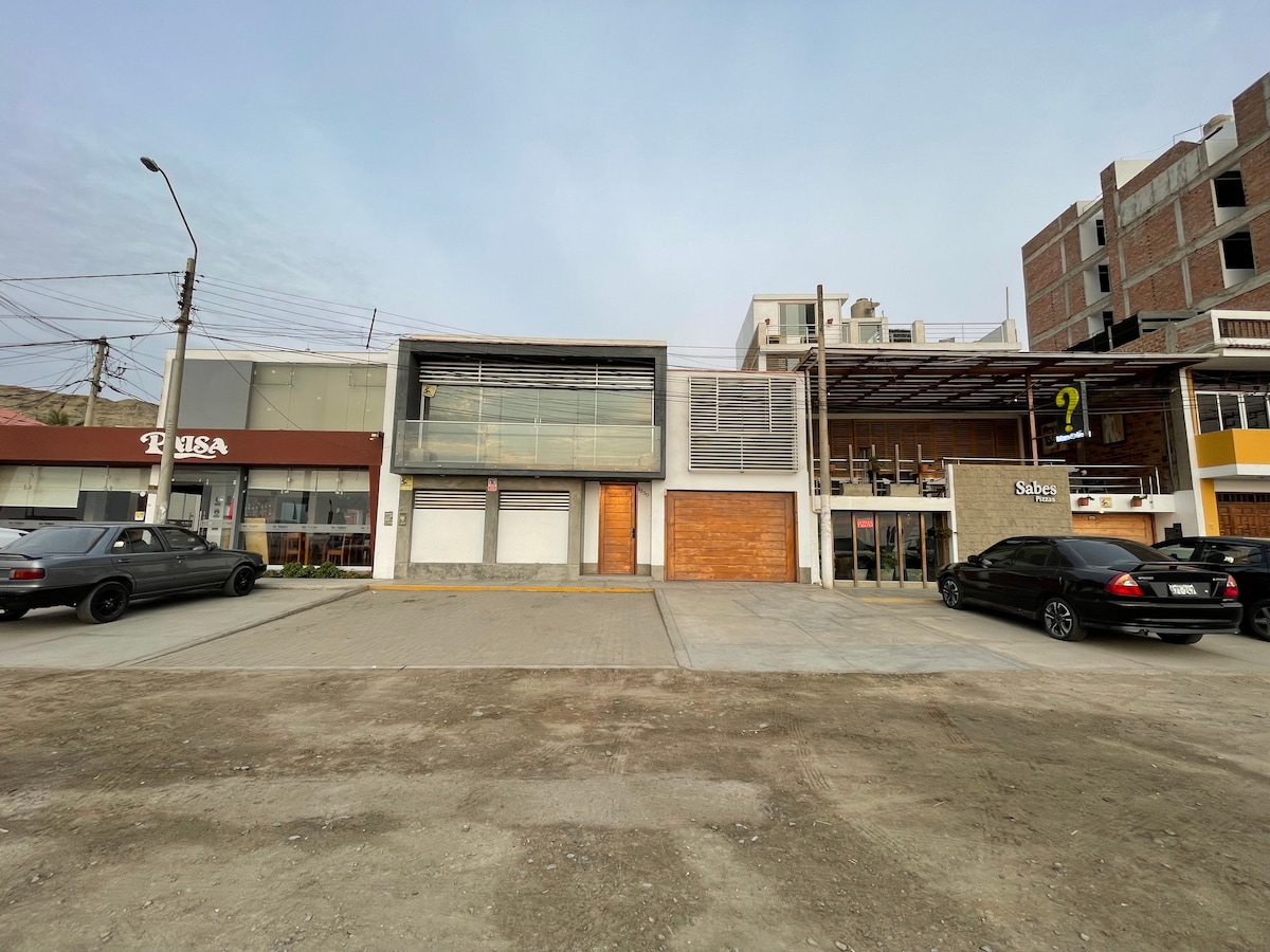 A modern two-story building is shown, featuring large glass windows and wooden doors. Adjacent commercial spaces are visible, with a combination of businesses occupying the ground level. The area is accessible with parking available for multiple vehicles, highlighting a communal and inviting atmosphere.