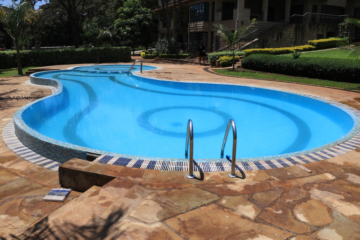 A curved swimming pool is outlined with patterned tiles, featuring clear blue water. Metal handrails provide access to the pool, surrounded by well-maintained gardens and lush greenery. sun loungers are positioned nearby, inviting relaxation in a serene outdoor setting.