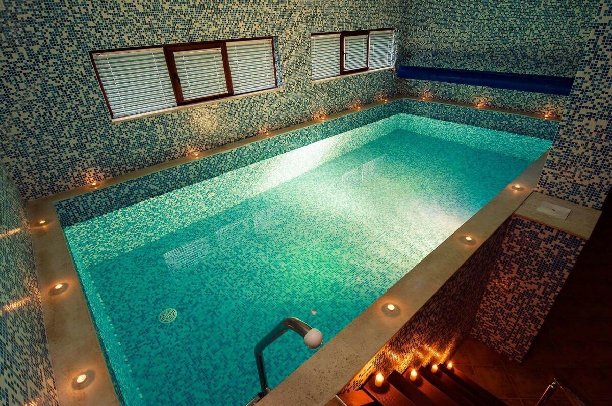 An indoor swimming pool features a bright blue tiled design, enhancing the tranquil atmosphere. Soft lighting surrounds the pool's edge, creating a serene ambiance. Large windows allow natural light to illuminate the space while providing a view of the outside.