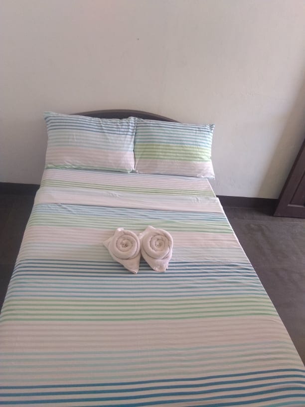 A simple sleeping area features a striped bedspread in soft blue and green tones atop a spacious bed. Two pillows rest at the head of the bed, and two rolled towels are arranged in a decorative manner.