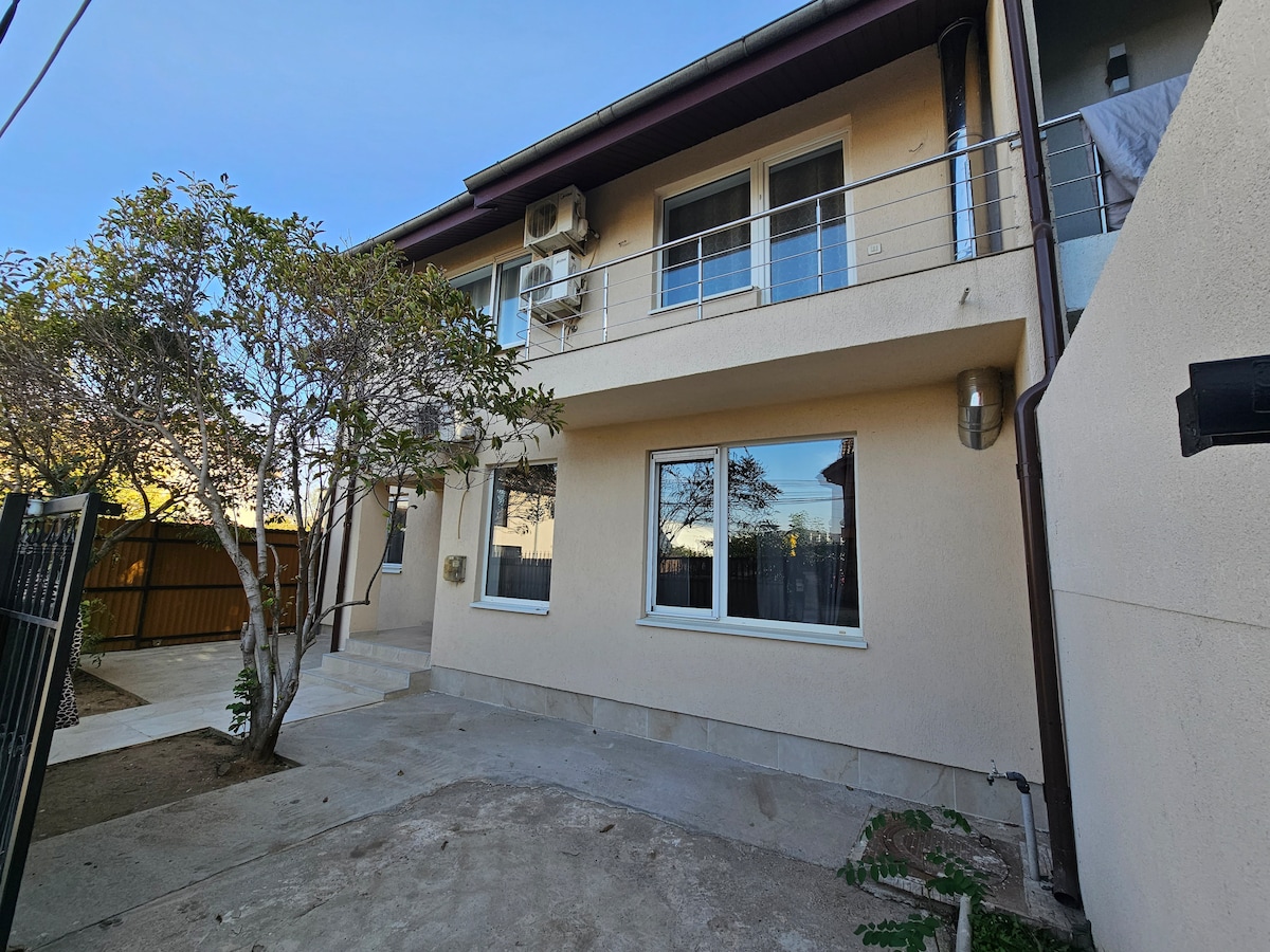 The exterior of a two-story building is shown, featuring large windows and a small outdoor area with gravel. A tree is positioned close to the structure, and there are air conditioning units mounted on the walls. A fence partially surrounds the property.