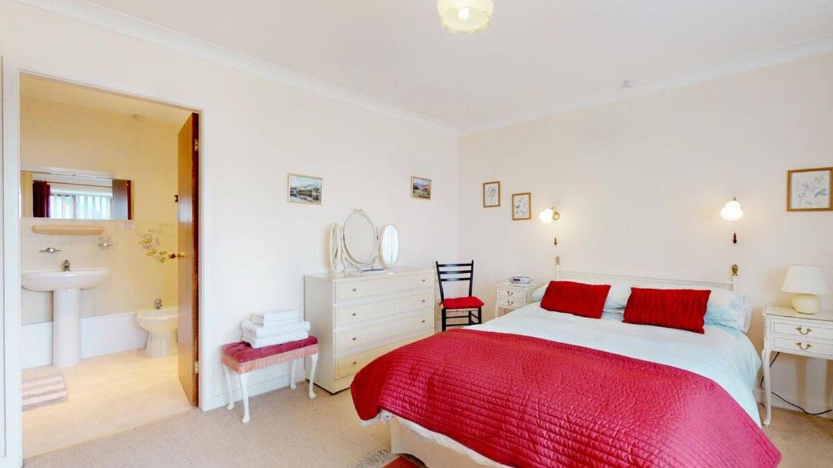 A well-appointed bedroom is presented with a spacious bed dressed in light bedding and red accents. A vanity table and chair are visible, along with a dresser and a welcoming armchair. An ensuite bathroom is accessible, featuring modern fixtures in a neutral colour palette.