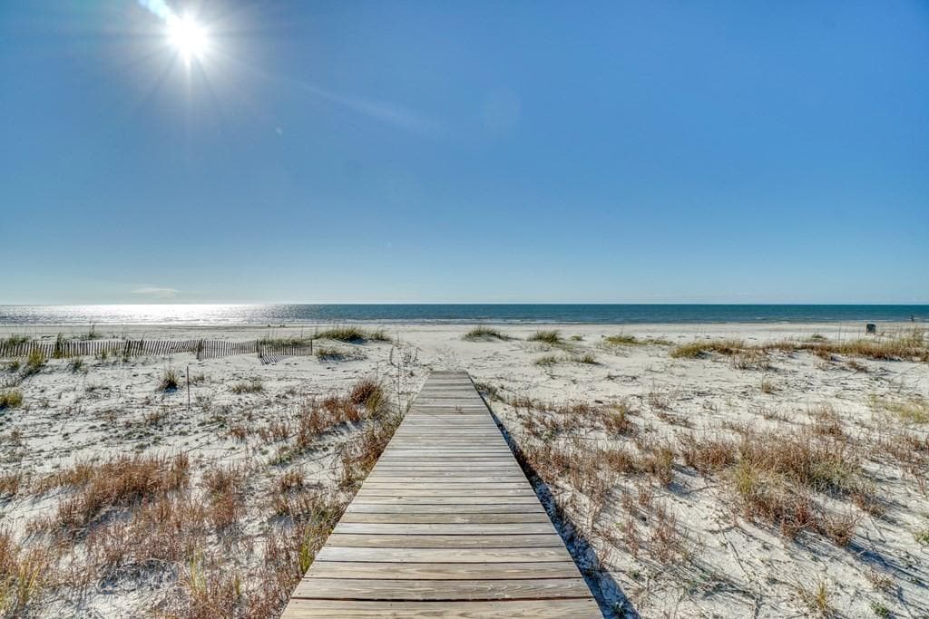A wooden boardwalk leads to a private beach, with white sand and sparse vegetation visible along the edges. Gentle waves can be seen in the distance under a clear blue sky, illuminated by sunlight reflecting off the ocean surface.
