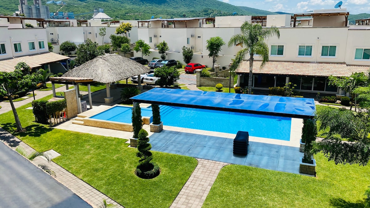 An outdoor area features a spacious blue swimming pool alongside a separate shallow children's pool. A shaded lounge area with a thatched roof is visible. Well-maintained gardens showcase neatly trimmed shrubs and greenery, contributing to a relaxed environment within the community.