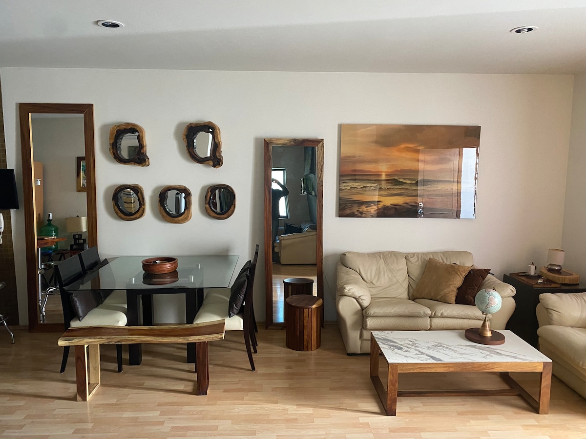 A living area is presented with a glass-top dining table surrounded by five dark chairs. A cream-colored leather sofa invites relaxation, while a wooden coffee table adds warmth. Reflections are seen in decorative mirrors on the wall, and artwork depicting a serene landscape adorns the space.
