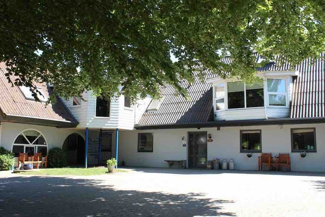 A welcoming exterior of the guest accommodations showcases a spacious building with a sloped roof and large windows. Overhanging trees provide shade to the entrance area, which features outdoor seating arrangements and decorative elements at the front.