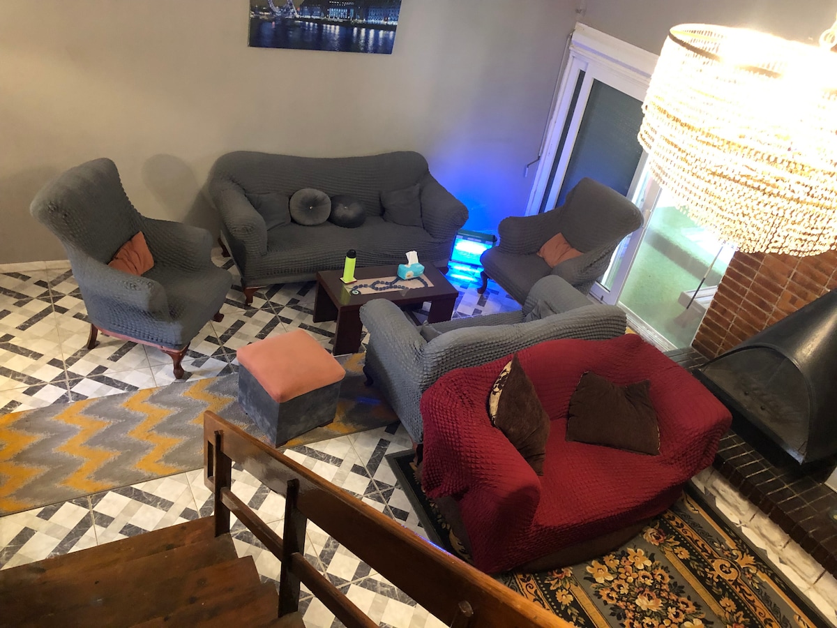 The living area features gray woven sofas, offering comfortable seating around a central coffee table. A modern chandelier provides overhead lighting, while a decorative rug adds warmth to the patterned tile floor. A staircase leads to an upper level, enhancing the room's spacious feel.
