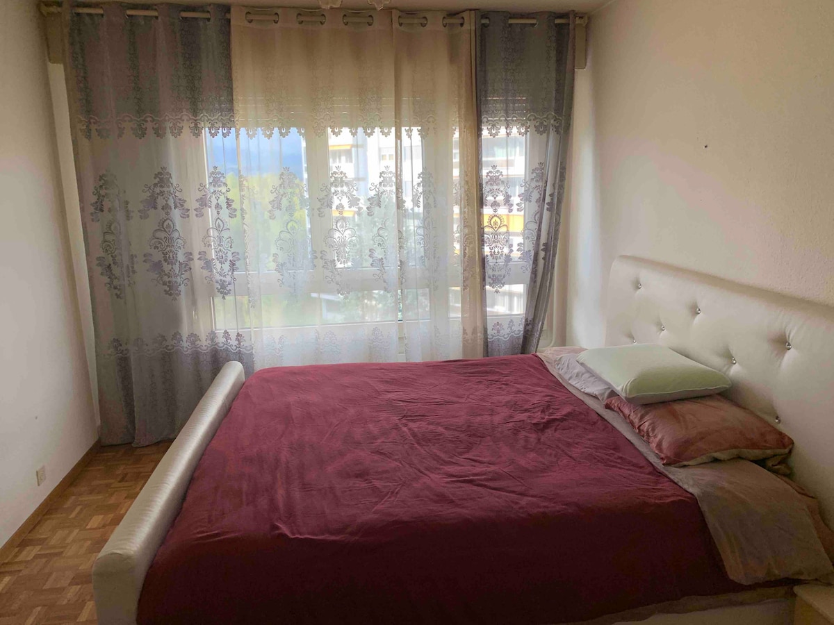 A spacious bedroom featuring a king-size bed with a red bedspread. Pillows are arranged on one side of the bed. Sheer curtains with floral designs allow natural light to filter in, providing a soft ambiance. A window is visible in the background, framing an outdoor view.