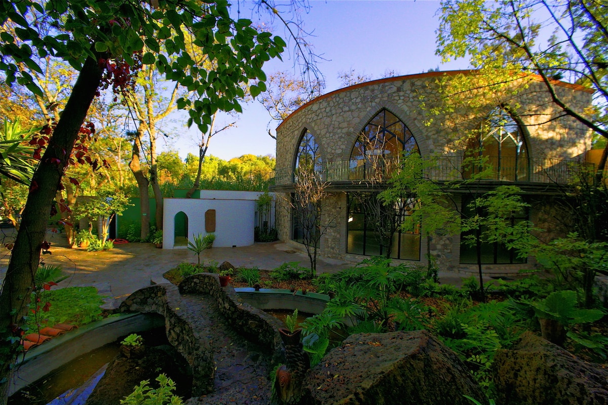 A unique stone building with large arched windows is framed by lush greenery and vibrant plants. A serene water feature flows through the garden, reflecting the surrounding natural beauty in a tranquil setting. The landscape features a harmonious blend of architectural and organic elements.
