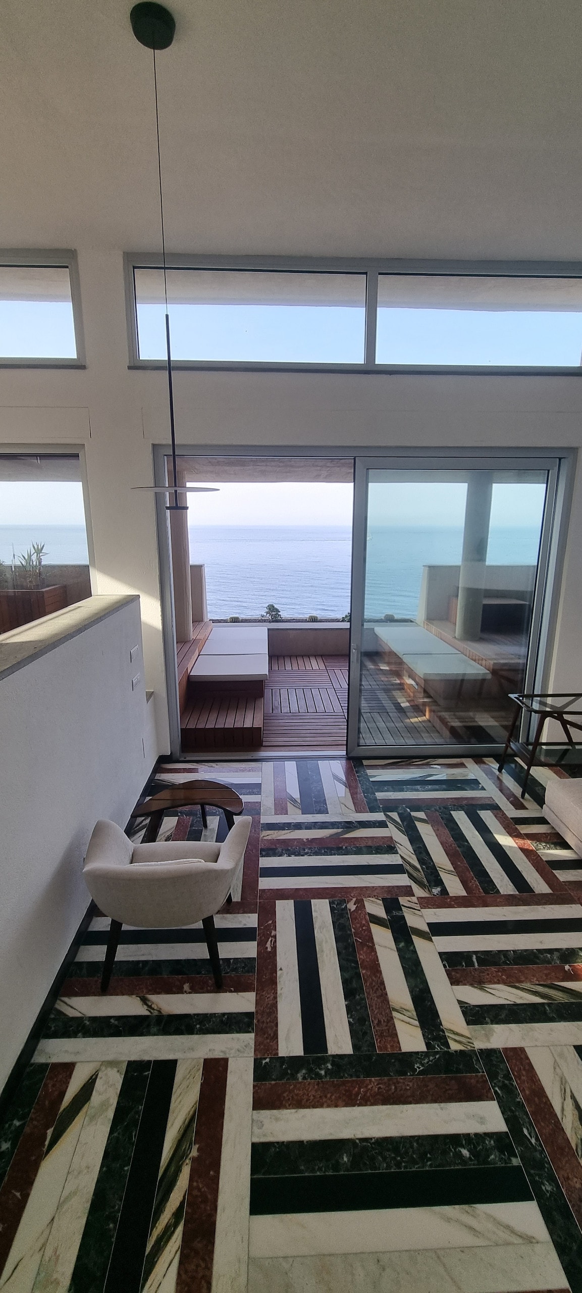 A spacious interior space features a striking marble floor with a geometric pattern. Large glass doors open to a panoramic view of the sea, allowing natural light to enhance the area. A comfortable chair and wooden seating can be seen near the window.