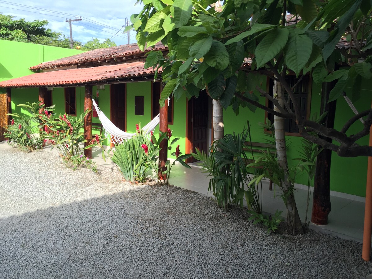 The vibrant green exterior of the property is complemented by a gravel pathway and lush tropical plants. A hammock is nestled between two wooden posts, inviting relaxation in a serene outdoor space. The layout blends traditional architecture with a natural environment.