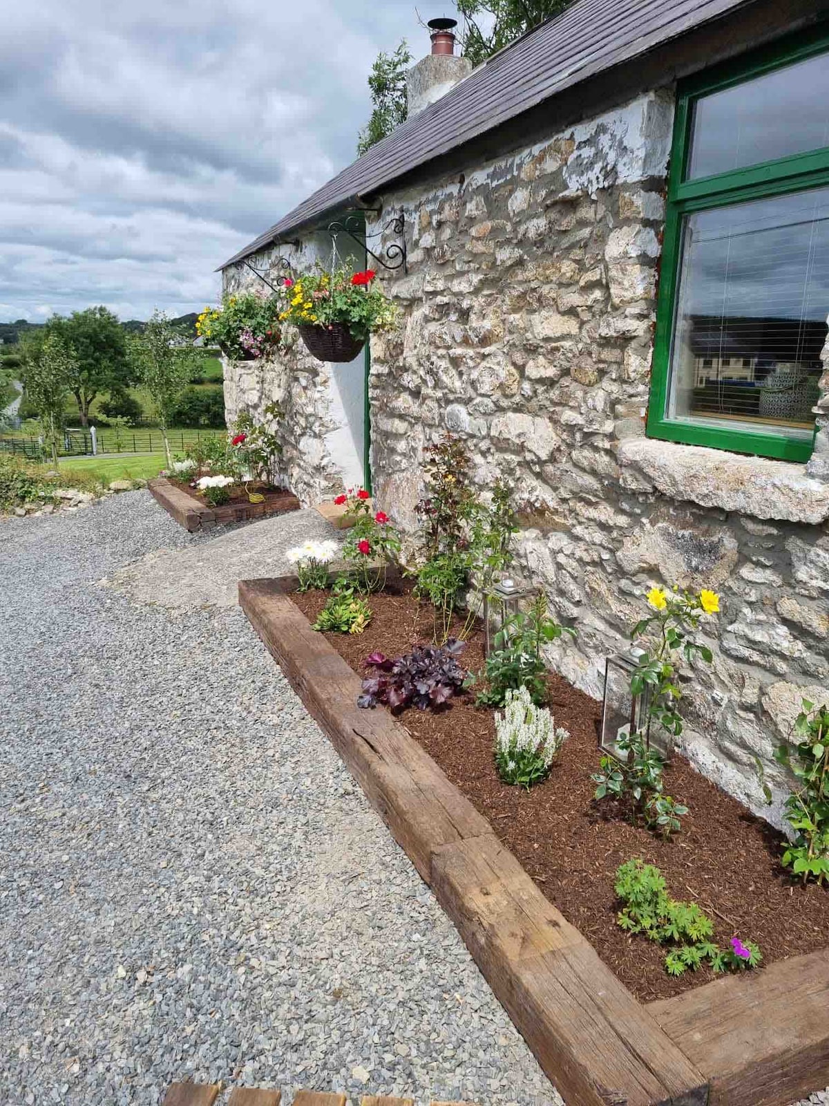 The exterior of the cottage features a stone facade with green trim. Flower beds are lined along the sides, showcasing a variety of colorful flowers and plants. A hanging basket filled with blooms adds charm to the entrance, complementing the tranquil outdoor setting.