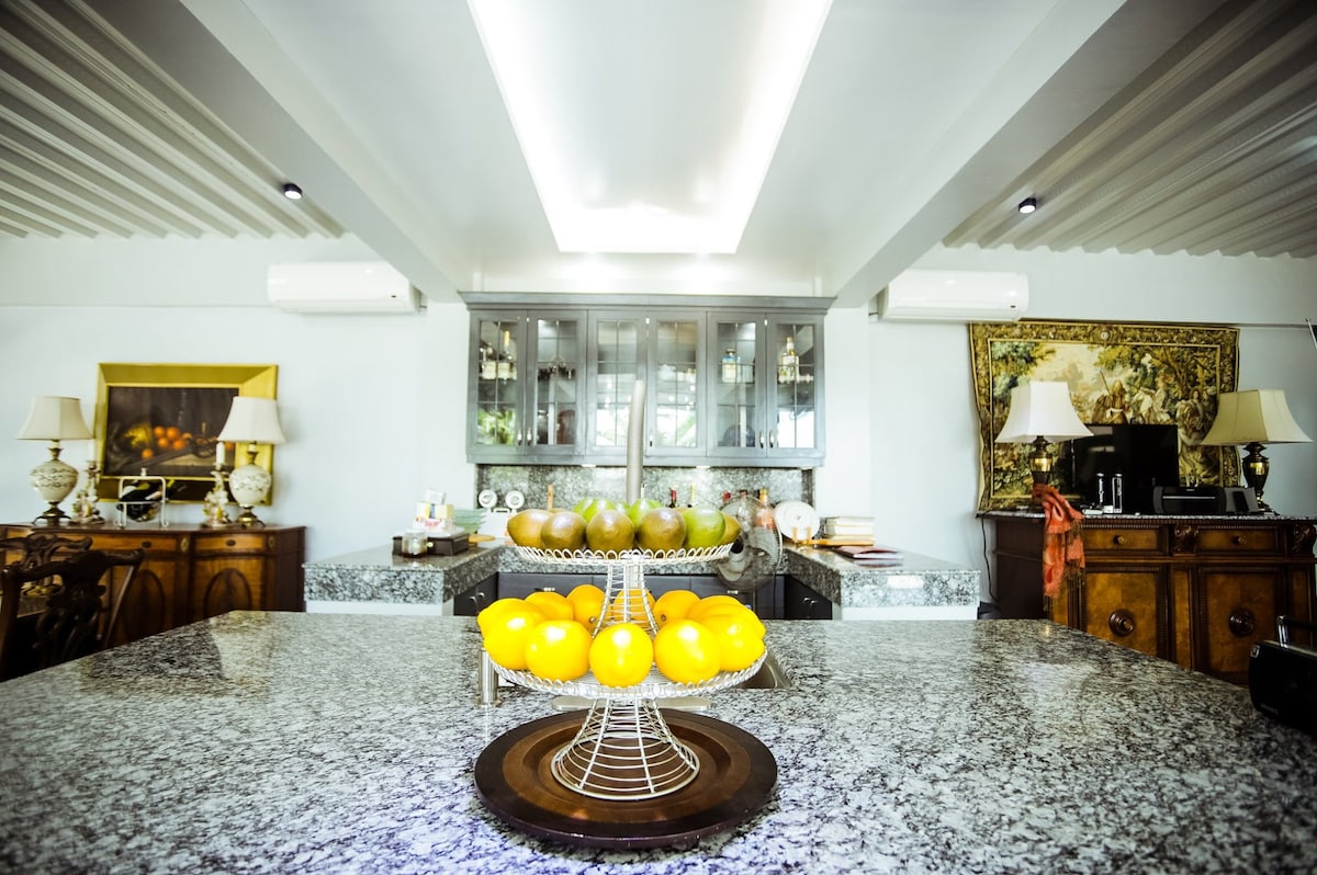 A modern kitchen features a sleek granite countertop with a tiered fruit stand holding vibrant lemons. Behind the counter, wooden cabinetry displays a variety of fresh produce. Soft lighting and decorative lamps create a welcoming ambiance, enhancing the functional layout of the space.