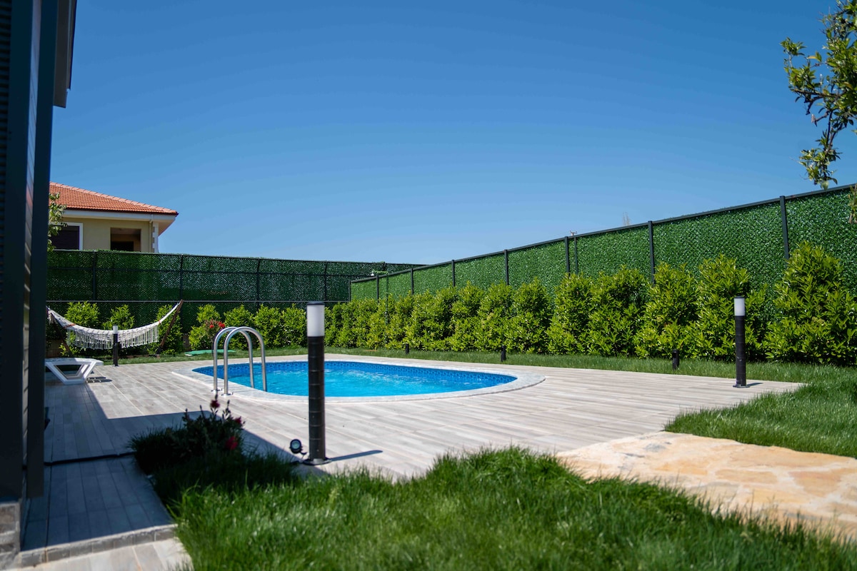 A serene outdoor space features a swimming pool surrounded by a wooden deck. Lush greenery lines the perimeter, and a hammock is positioned between two posts. Modern lighting fixtures are placed along the edge of the deck, enhancing the tranquil atmosphere.