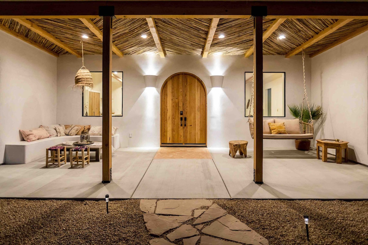 The entrance features a wooden door framed by two large windows, providing natural light. A shaded porch area is furnished with a comfortable lounge area, a wooden table, and a swing chair. Decorative plants and soft lighting enhance the welcoming atmosphere.
