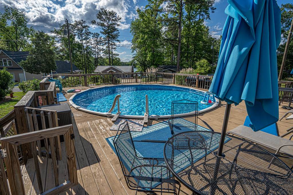 A private pool is featured on a wooden deck, surrounded by a railing and equipped with lounge chairs and a shaded umbrella. Clear water reflects the sky, with lush greenery in the background providing a serene environment.