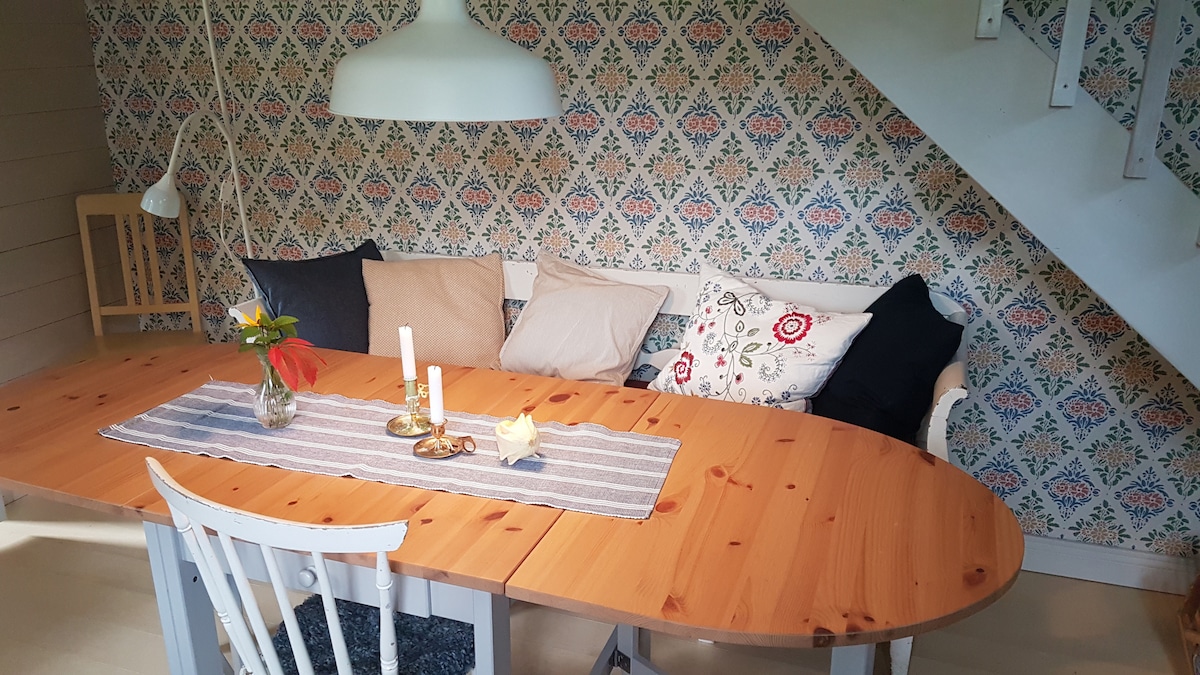 A dining area is presented with an oval wooden table at the center, surrounded by white chairs. A patterned wallpaper adds color to the space. A cozy seating arrangement features cushions in various patterns and colors. A vase with flowers and candles enhance the table setting.