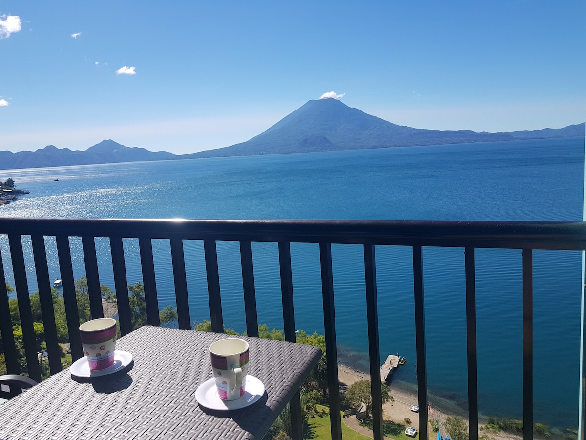 A balcony with a dark wicker table is showcased, holding two cups positioned neatly. In the foreground, the calm blue waters reflect sunlight, while majestic mountains rise in the background against a clear blue sky.