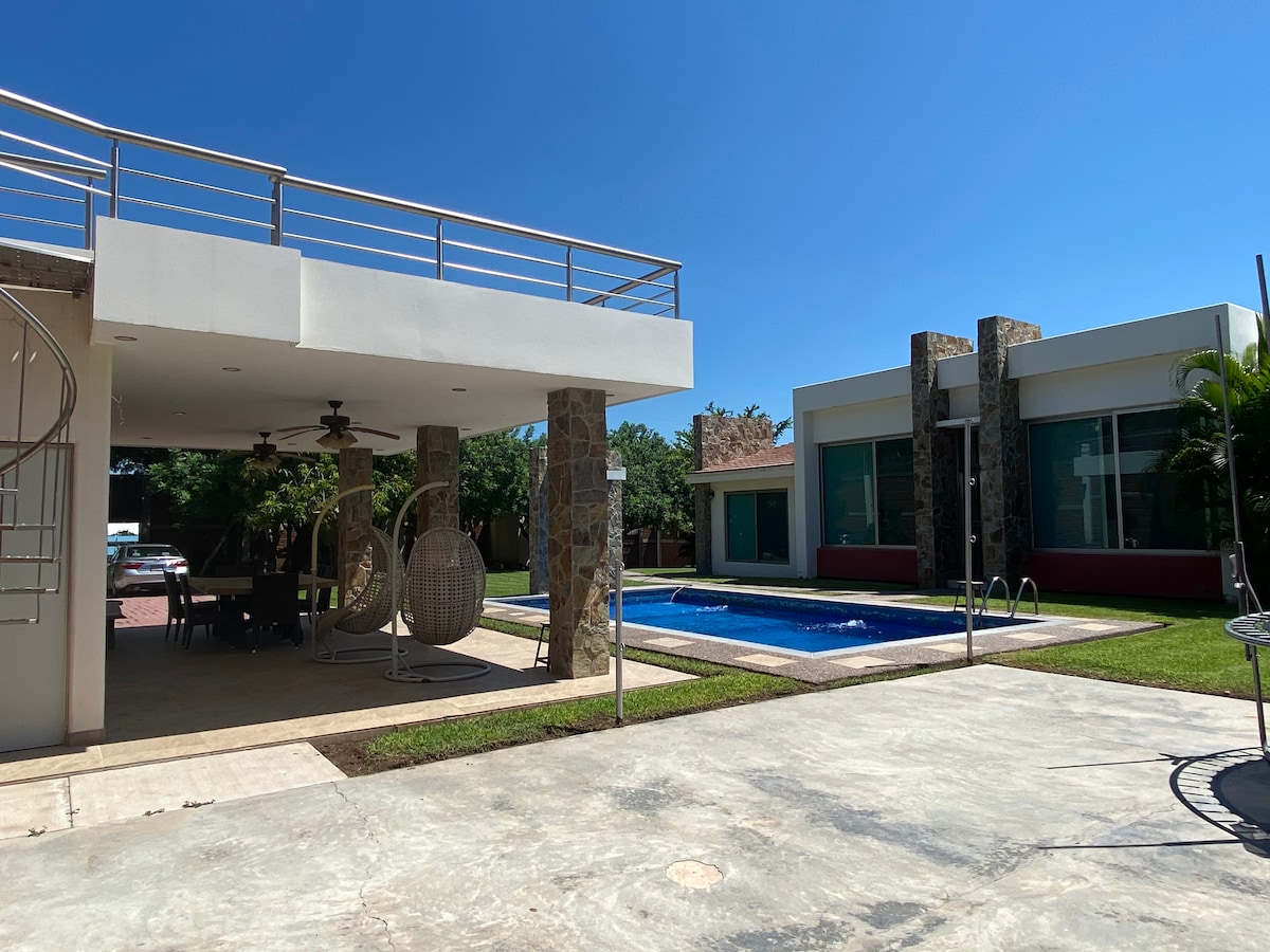 A modern outdoor space features a swimming pool surrounded by greenery. The house, with large windows, showcases a clean architectural design. A covered patio, equipped with hanging chairs and ceiling fans, is visible, enhancing the area for relaxation. Clear blue skies provide a bright backdrop.