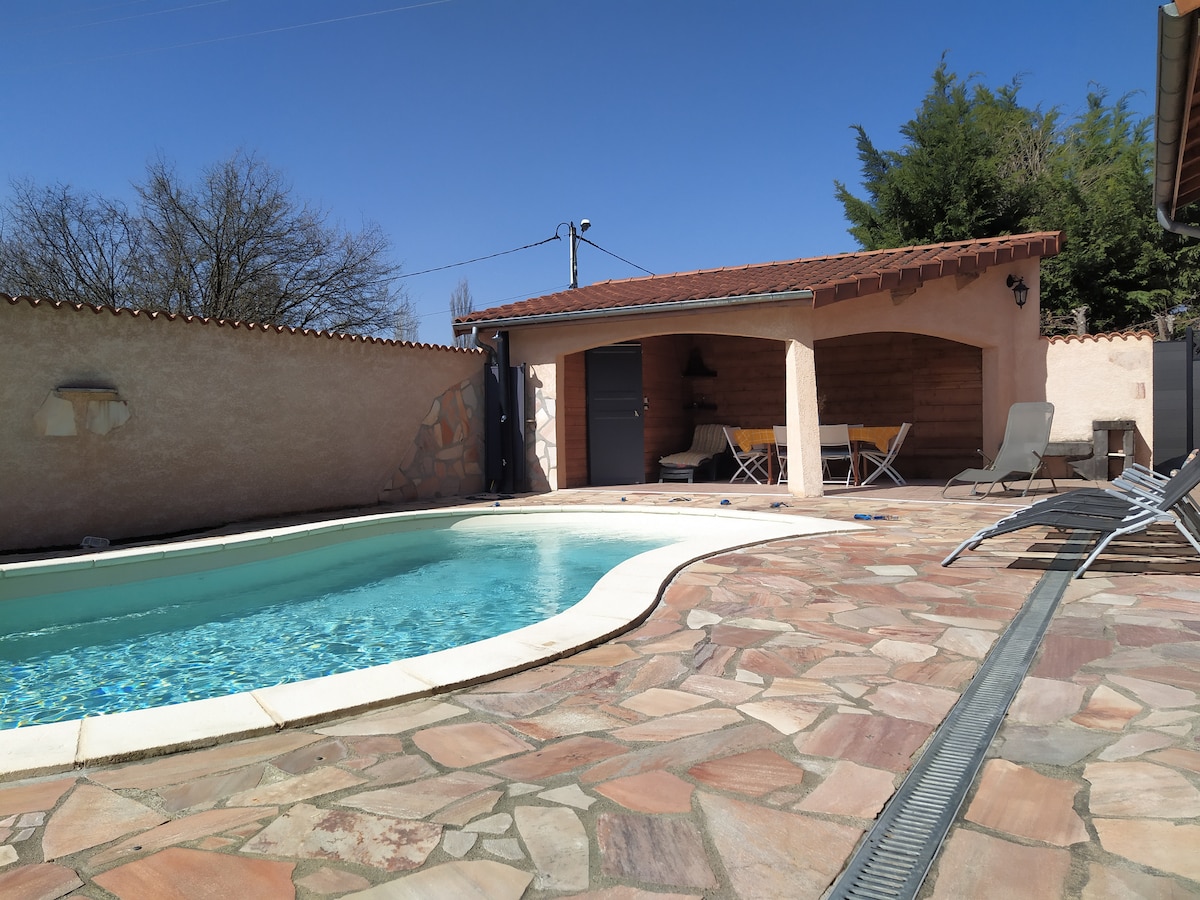 A pool area is outlined by charming stone paving. The pool shines in sunlight, with a covered terrace featuring a dining table and chairs ready for gatherings. Lounge chairs are arranged nearby under a clear blue sky, providing a relaxing environment.