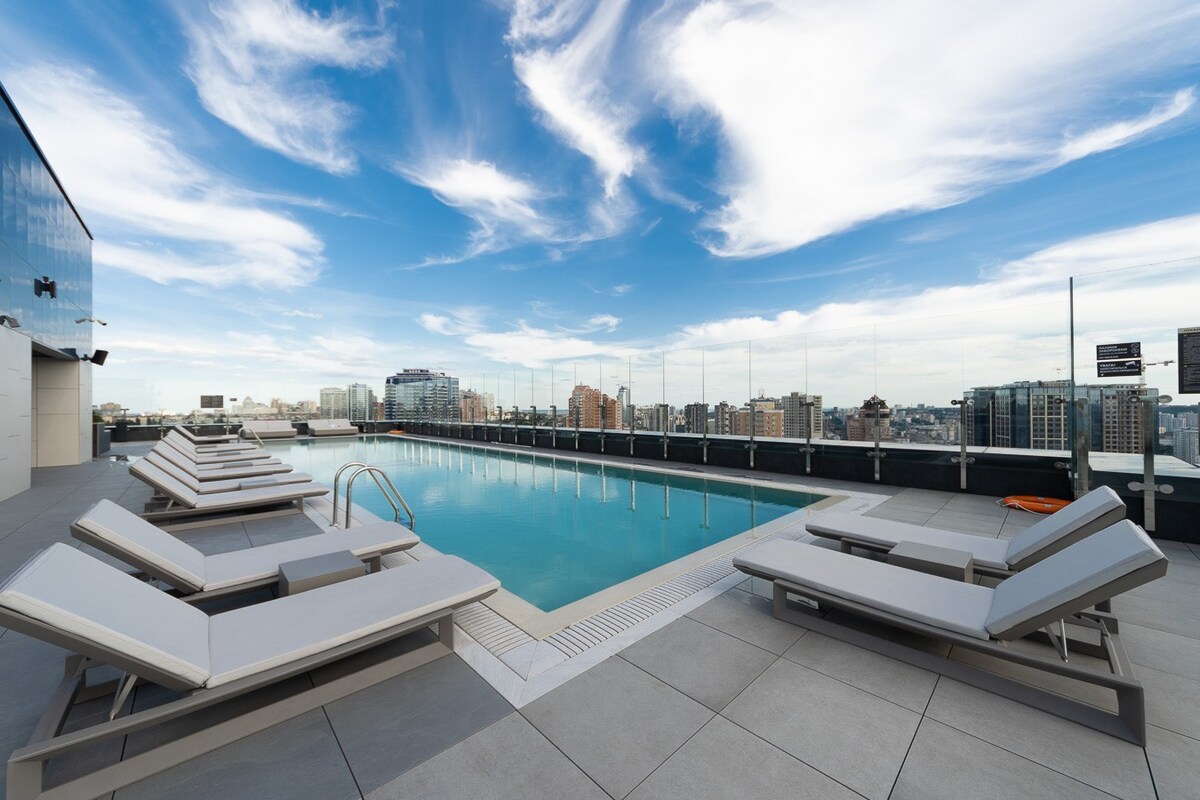 A rooftop area features a wide swimming pool surrounded by multiple lounge chairs. The city skyline is visible in the background under a clear blue sky with scattered clouds. The pool's edge is lined with a smooth deck, offering a relaxing atmosphere.