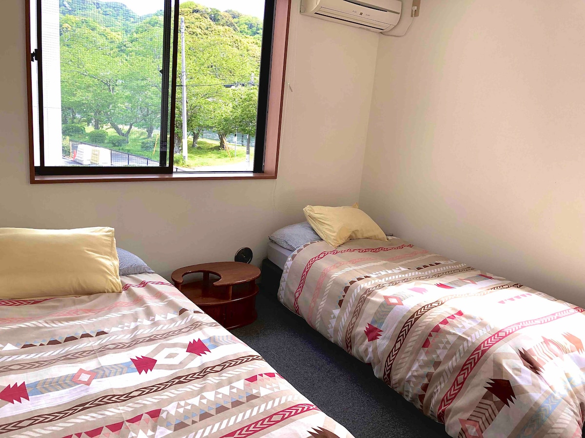 A bright room is furnished with two beds, each dressed in patterned bedding. Soft pillows are placed on the beds. A small table is positioned between them. Natural light enters through a large window overlooking greenery outside.