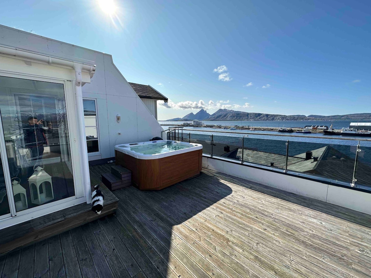 A private terrace features a wooden deck and a spacious Jacuzzi, surrounded by scenic views of the water and mountains in the distance. Glass sliding doors lead into indoor spaces, while sunlight illuminates the area, creating an inviting outdoor atmosphere.