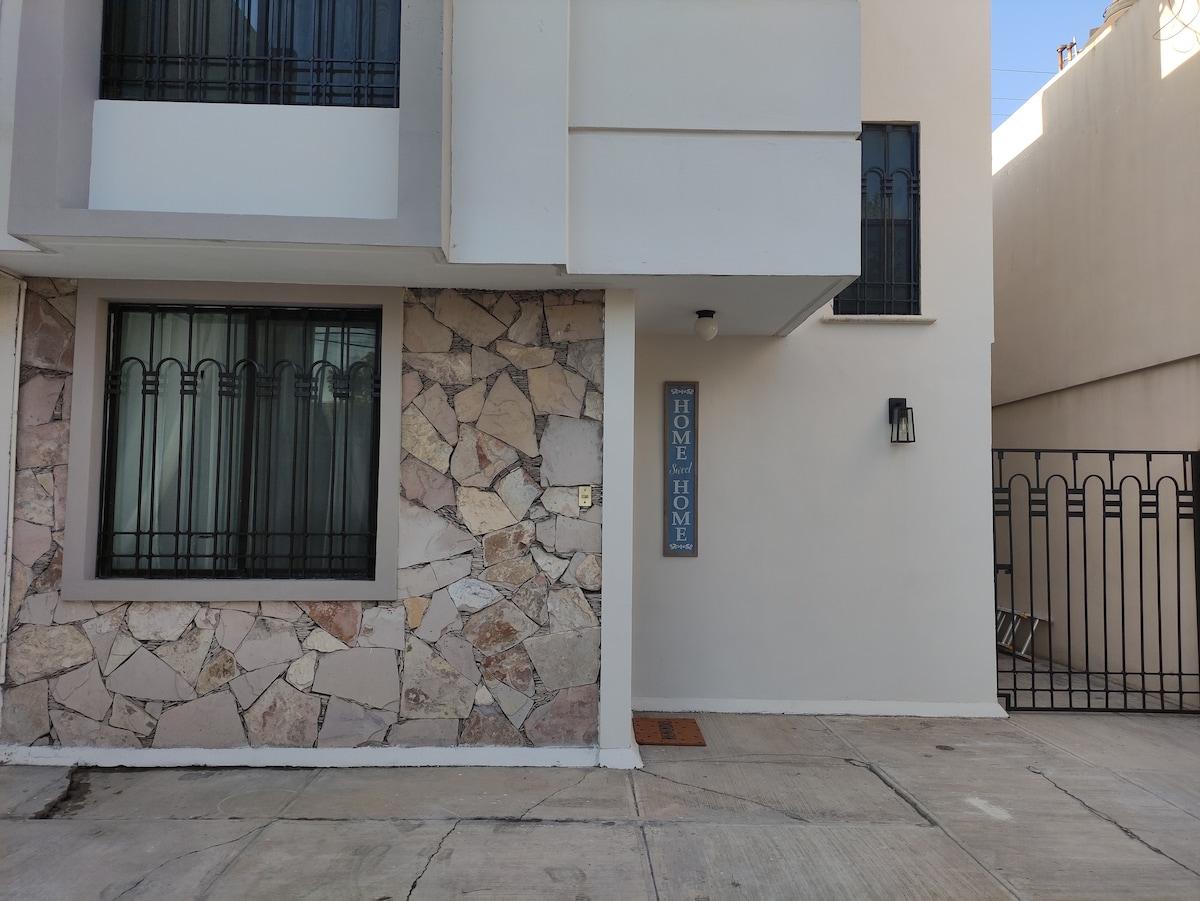 A modern entryway is depicted, featuring a blend of light-colored stucco and stone accents. A sign reading 'HOME' is prominently displayed. A window with decorative black bars allows natural light, and a gated entrance is visible in the background.