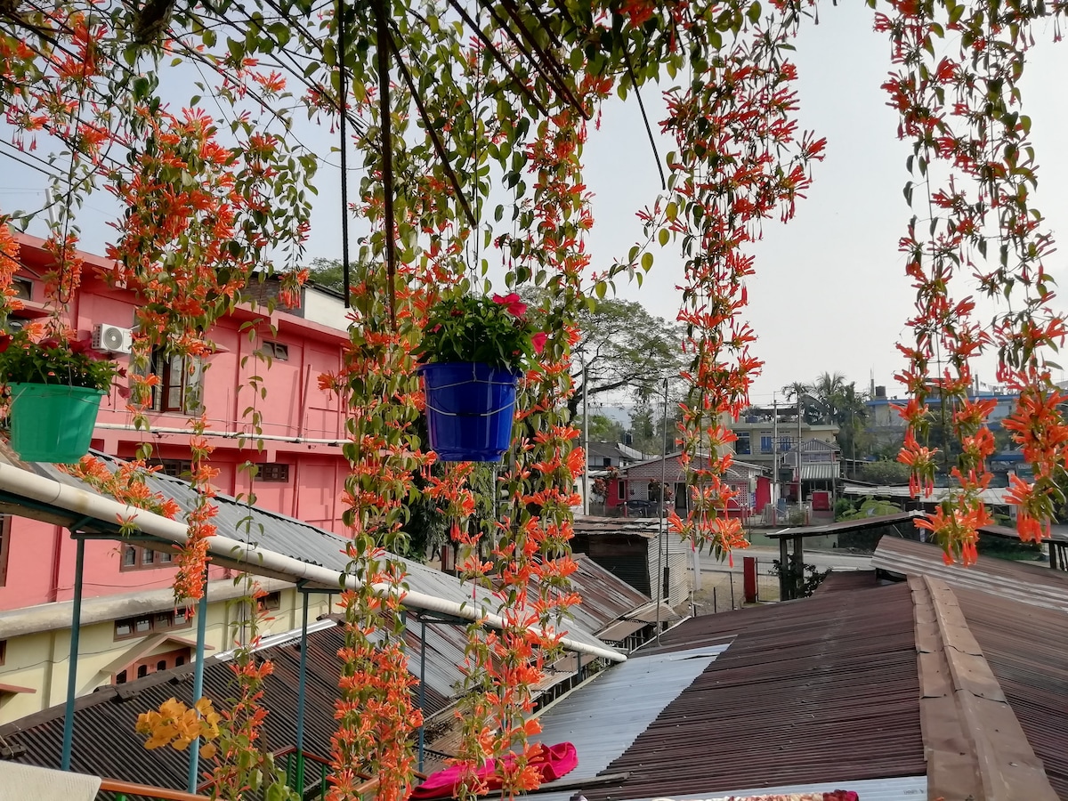 Hanging flower pots filled with vibrant orange blooms add a touch of color to the outdoor space. An umbrella provides shade, surrounded by rooftops and greenery in the village. The scene reflects a blend of nature and local architecture.