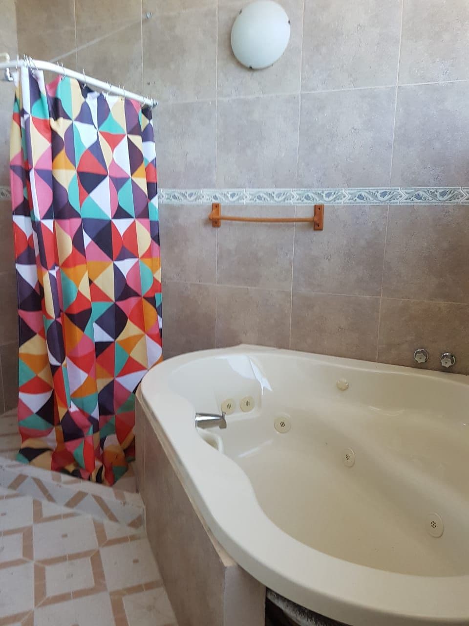 A spacious bathroom features a modern corner bathtub surrounded by neutral tiled walls. A colorful geometric shower curtain adds a vibrant touch. A wooden towel rack is mounted on the wall, and natural light is provided by an overhead light fixture.