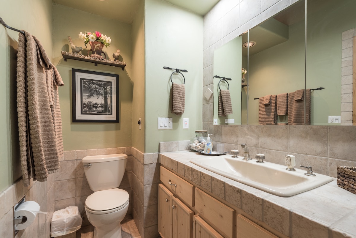 A clean and functional bathroom is displayed, featuring a double sink vanity with a stone countertop. A large mirror reflects the space, which is complemented by soothing green walls. Soft towels hang on racks, and decorative elements are arranged on a shelf above the sink.