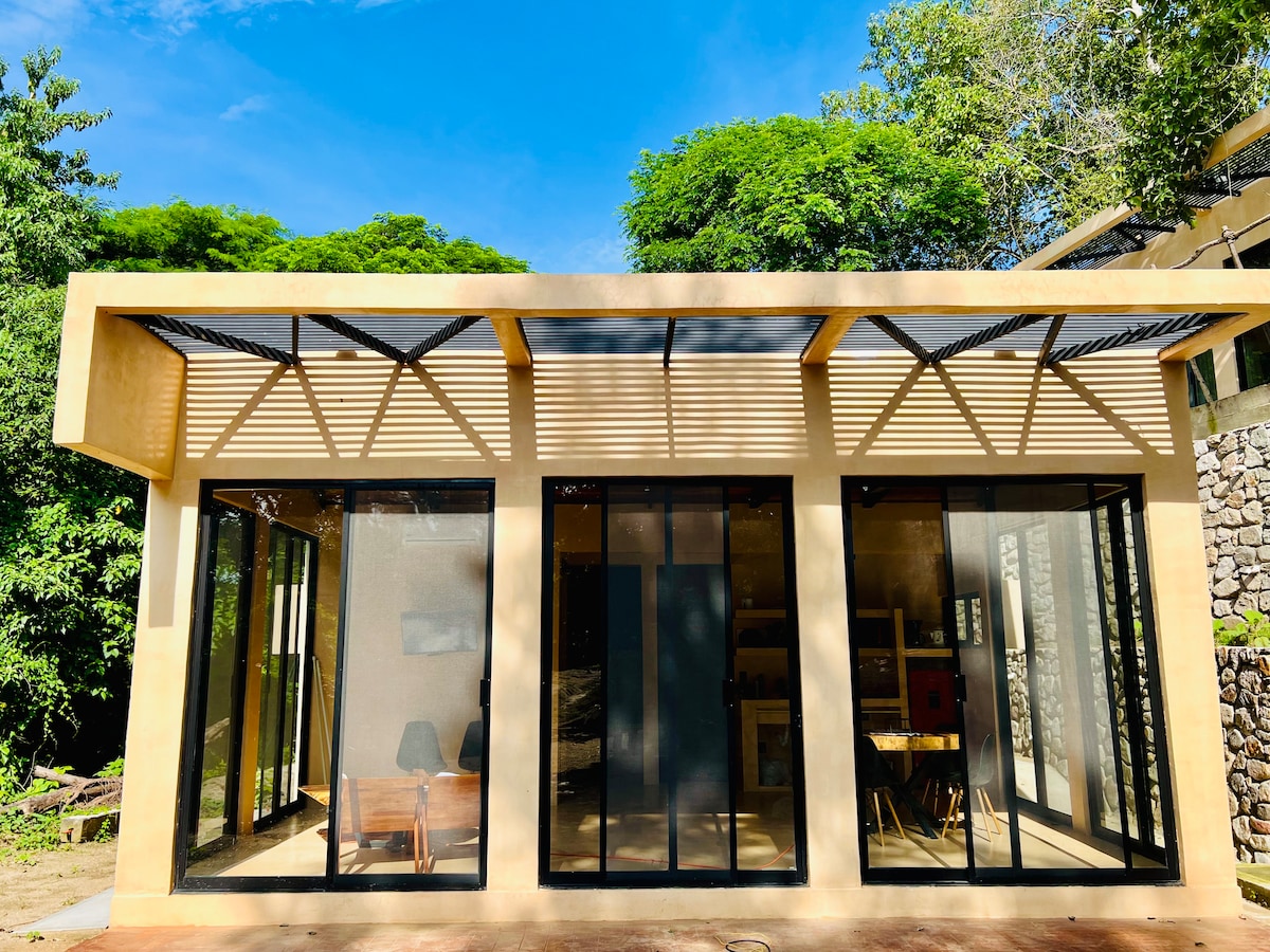 The exterior of a modern villa is presented, featuring large glass windows and a slatted roof providing shaded areas. Lush greenery surrounds the property, enhancing the connection with nature. The design emphasizes natural light and offers a clean, open appearance.