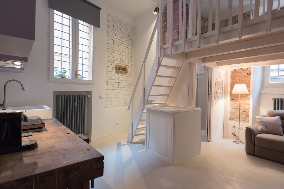 A bright living space showcases a mix of modern and rustic design elements. Exposed brick and whitewashed walls create a fresh ambiance. A cozy sofa is positioned next to a contemporary floor lamp. A staircase leads to an upper loft area, enhancing the spacious feel.