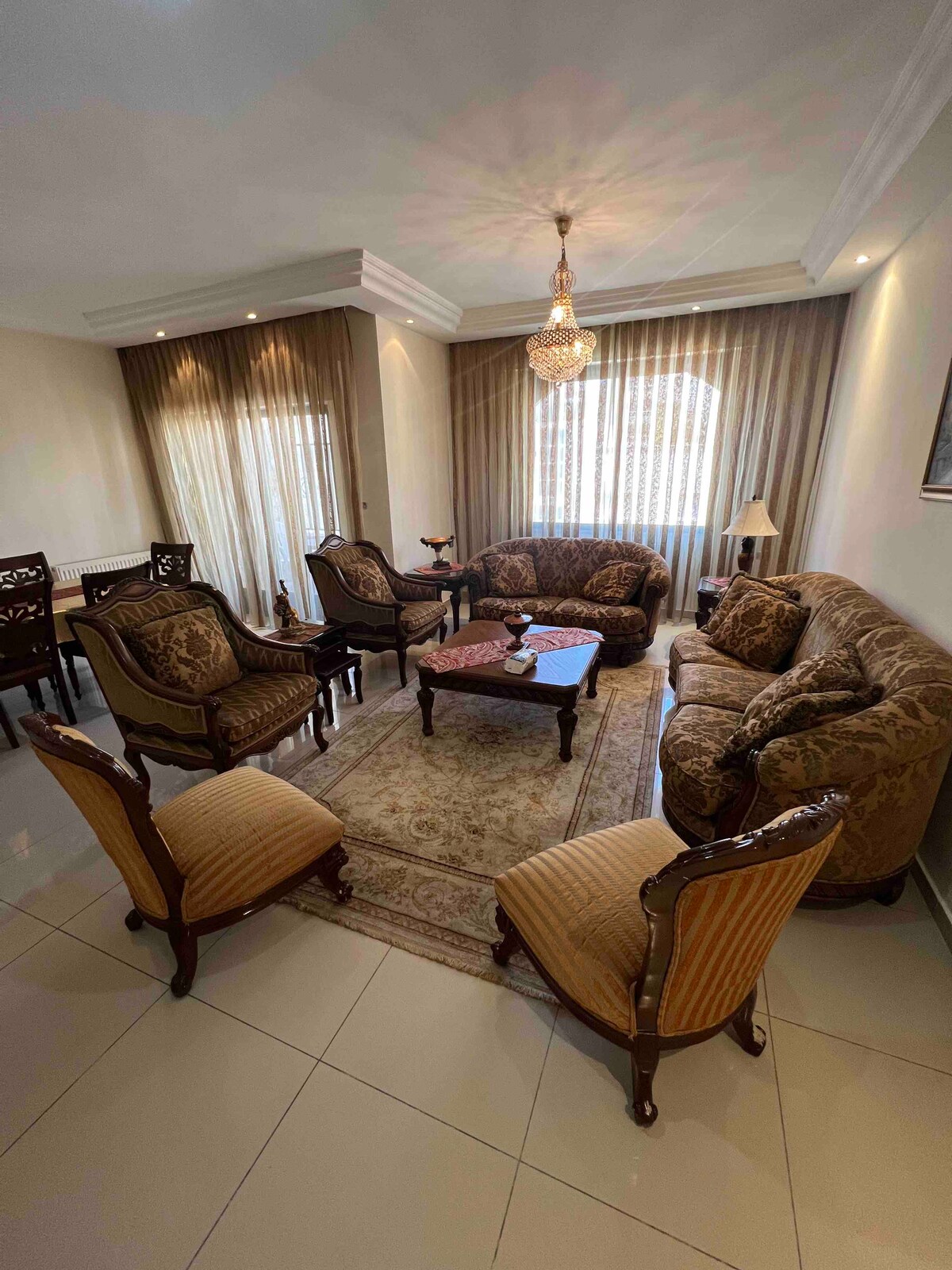 A spacious living room is furnished with an ornate seating arrangement, featuring plush sofas and patterned chairs. A decorative coffee table sits centrally on an area rug, and light filters through sheer curtains, enhancing the room's inviting ambiance.