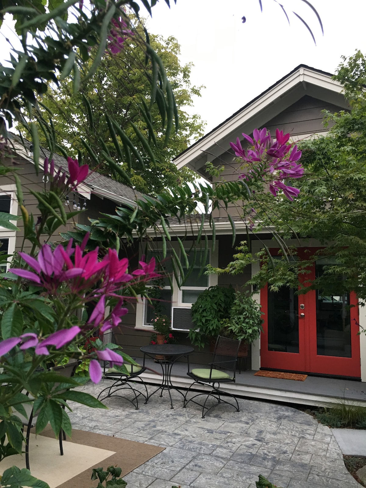 An inviting entrance is framed by vibrant purple flowers and lush greenery. The home's exterior features a warm, red front door and a beautifully designed pathway leading to a small seating area with a round table and chairs under the soft shade of the trees.