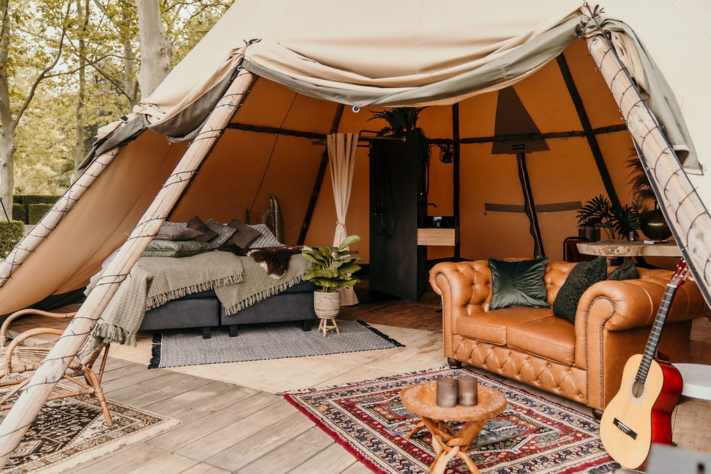 A comfortable interior of a spacious tipi includes a cozy Chesterfield sofa and a wooden table with chairs. A large bed is adorned with layered blankets, and houseplants add a touch of greenery. The entrance is framed by natural light filtering through the tent flaps.
