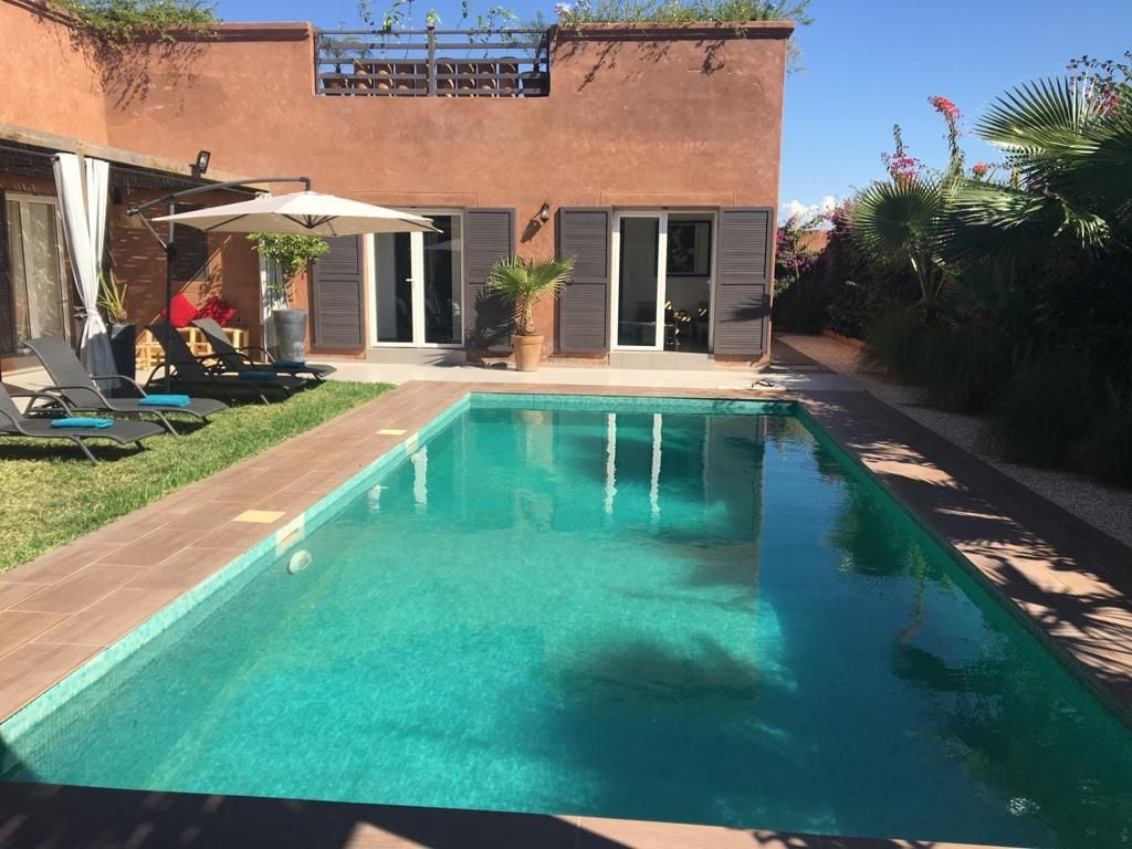 A private pool is framed by two rows of sun loungers, with a patio umbrella providing shade. The poolside is bordered by lush greenery and features a terracotta-colored villa with large sliding doors that open to the garden.