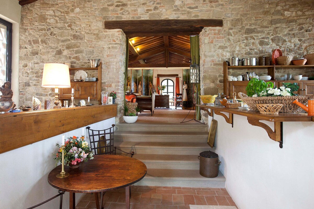 An inviting entryway leads to a spacious living area, featuring rustic stone walls and warm wooden accents. A small table adorned with flowers sits beside a staircase, with open shelves displaying various kitchen items and decor. Natural light enhances the welcoming ambiance.