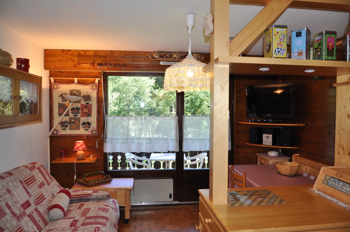 A cozy sitting area features a comfortable sofa and wooden accents, with sunlight filtering through large windows. A dining table and chairs are positioned nearby, and a decorative light hangs from the ceiling. A view of the outdoor space, adorned with a balcony, is visible.