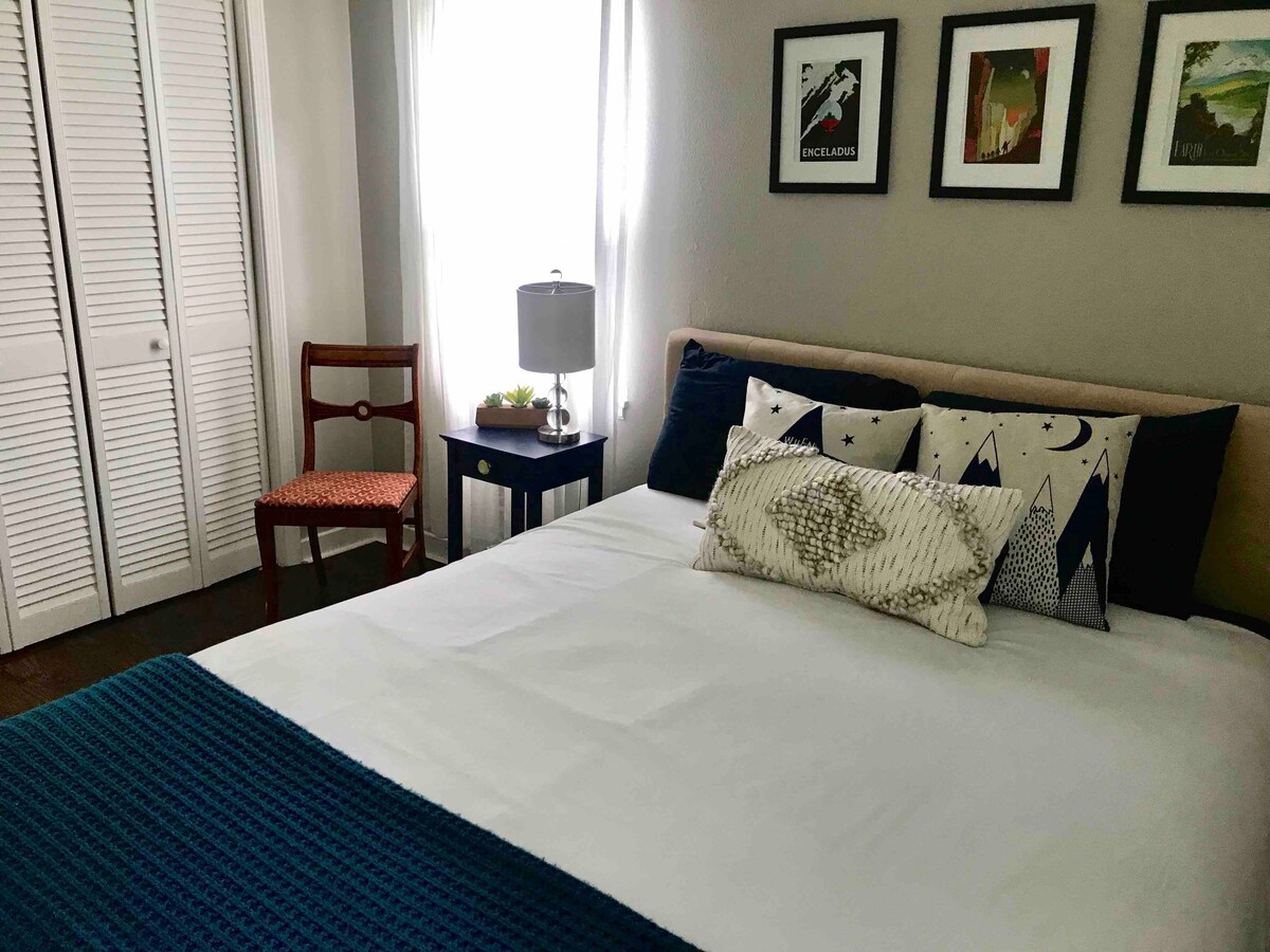 A cozy bedroom features a spacious bed with decorative pillows and a textured blanket. A bedside lamp rests on a small table alongside a decorative plant. Light filters through sheer curtains, complementing framed artworks on the wall and a wooden chair positioned nearby.