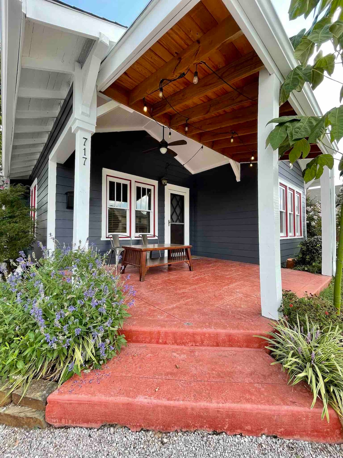 A welcoming front porch is presented with a covered area, featuring a wooden table and chairs. Lush flowering plants and greenery border the entrance, complemented by a gray-blue exterior and a rustic wooden ceiling. Warm lighting adds a cozy touch to the outdoor space.