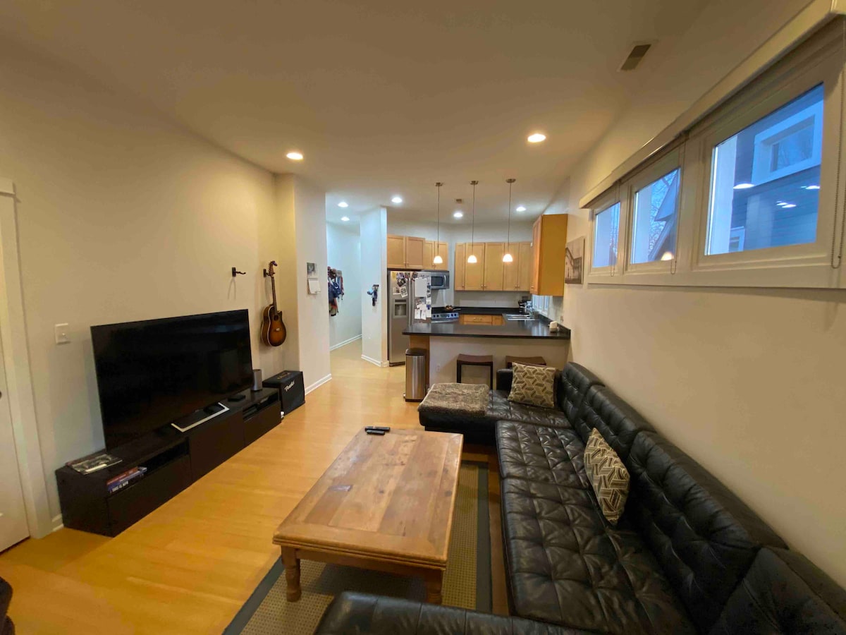 A spacious living area showcases a dark leather sectional sofa positioned near a wooden coffee table. A large flat-screen TV is mounted on the wall opposite the sofa. The open layout leads to a modern kitchen with warm wood cabinetry, illuminated by pendant lighting.